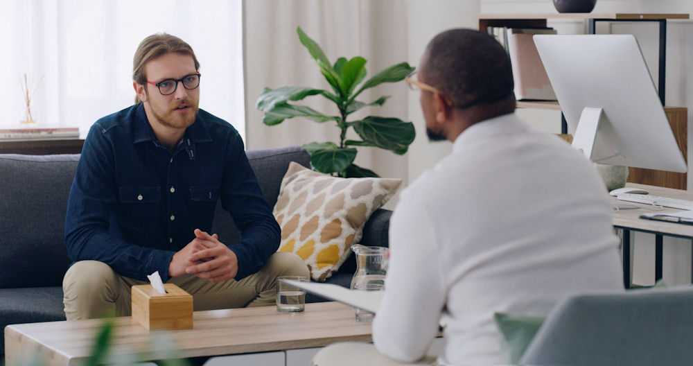 man wearing glasses and blue shirt sitting on a couch and speaking with counselor in php rehab